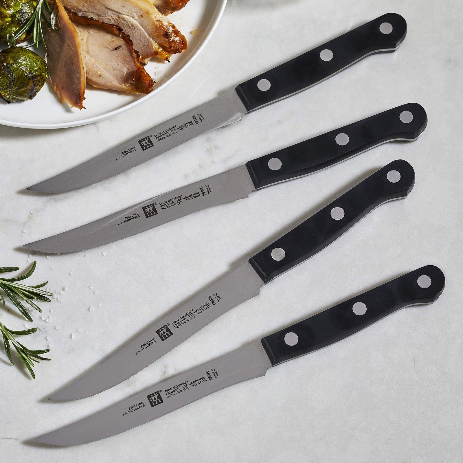 Set of four ZWILLING TWIN Gourmet steak knives with stainless steel blades and black handles, arranged on a wooden surface.