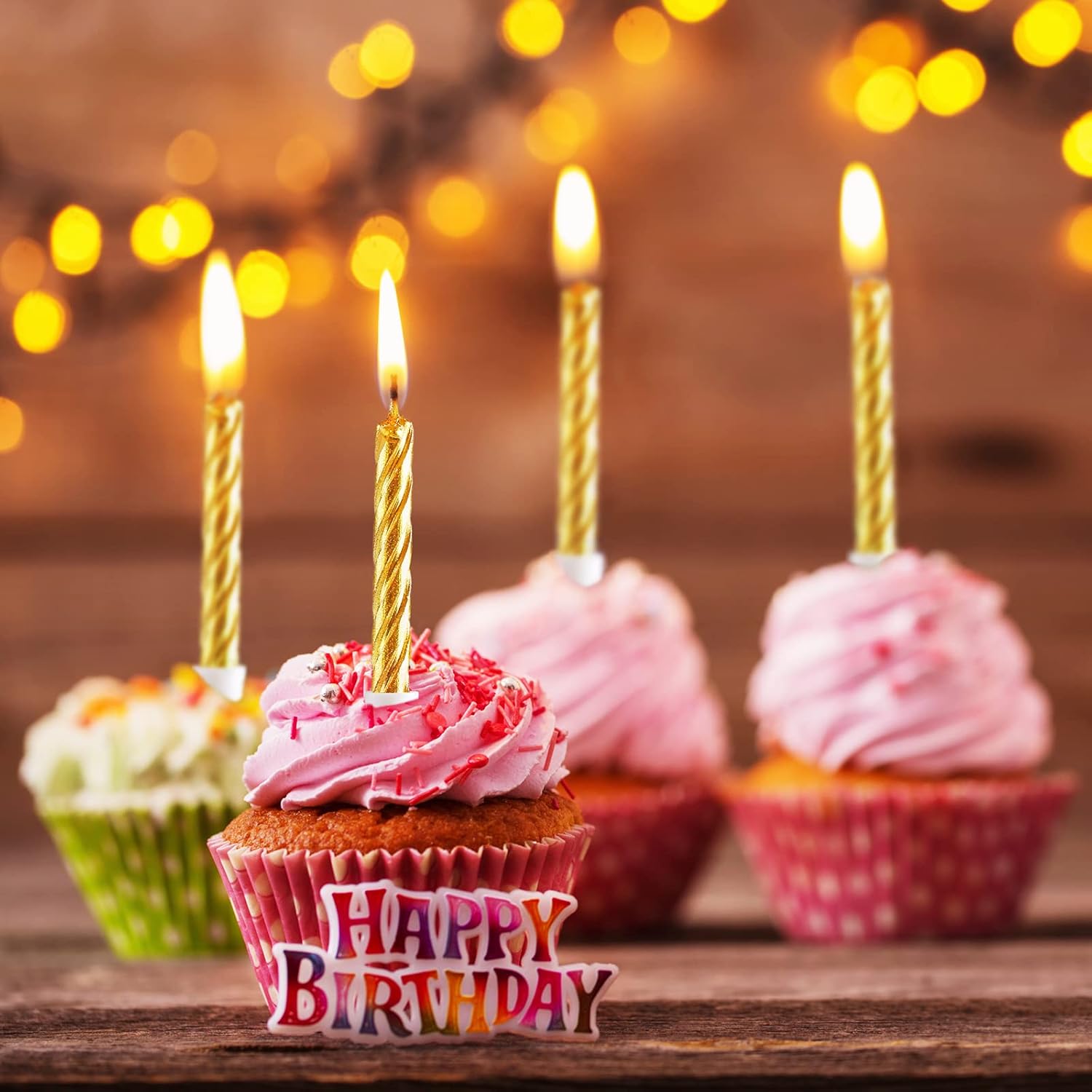 Birthday cupcakes with lit candles and a 'Happy Birthday' sign on a wooden table with blurred lights in the background.
