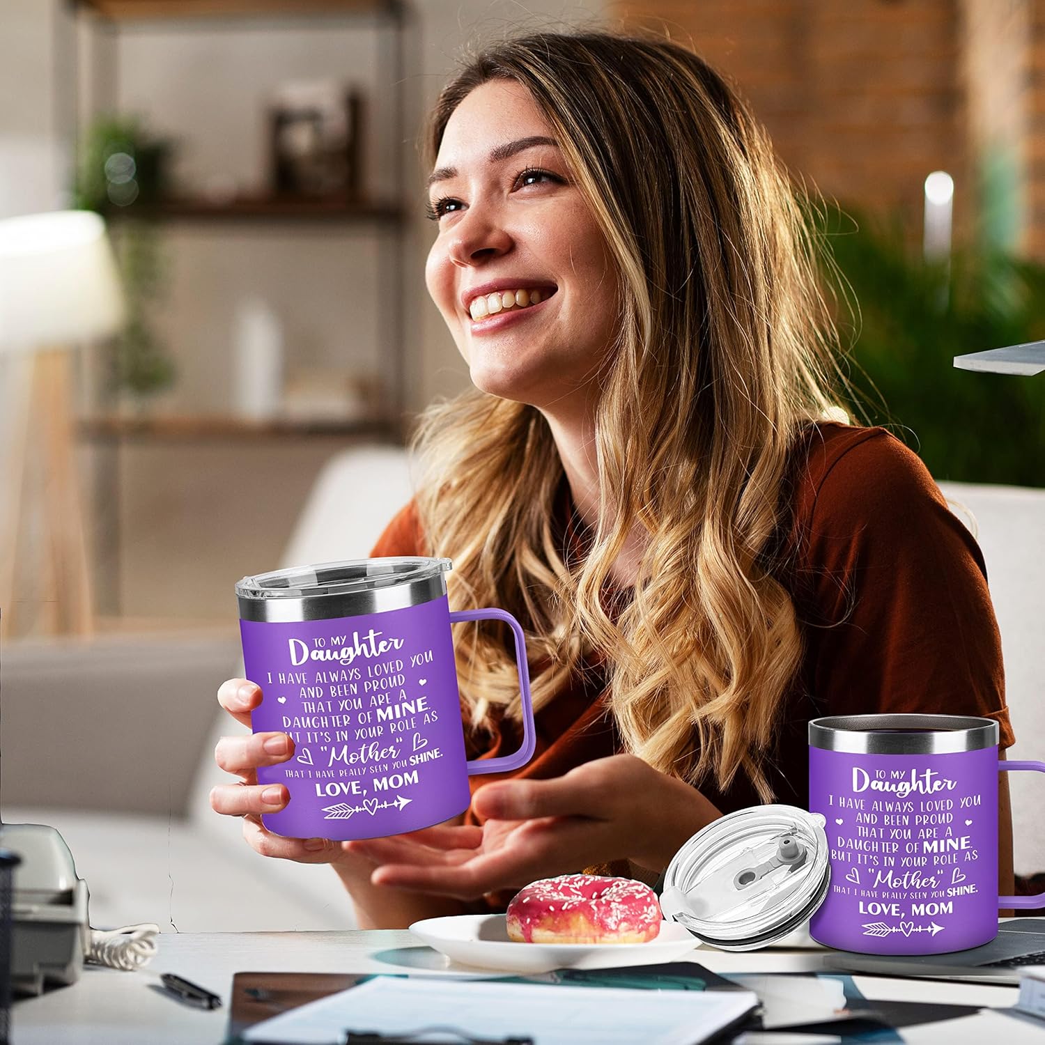Woman holding a purple mug with text, sitting at a table with a plate of donuts.